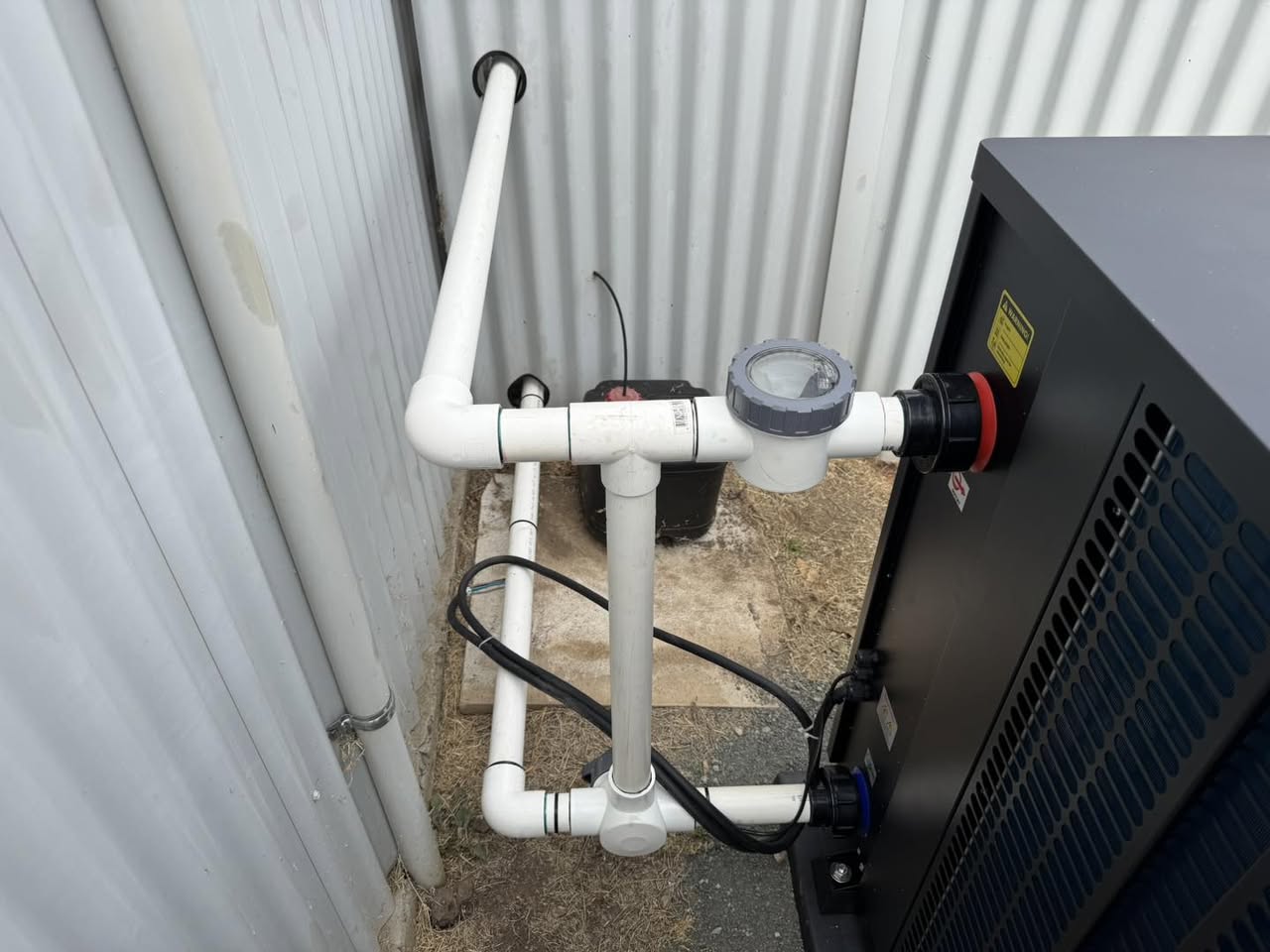 White PVC pipework with a sight glass and unions connected into the side of a dark inverter heat pump beside a shed wall.