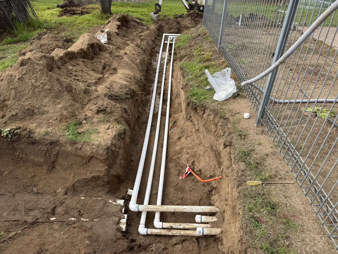 Four lengths of white PVC pipe laid along the bottom of an open trench in a backyard next to a fence.