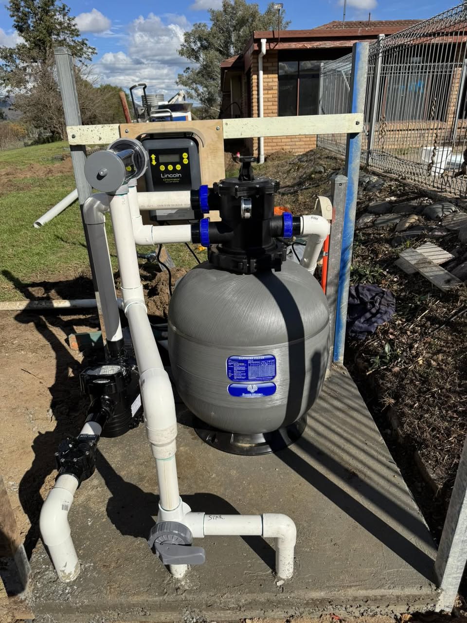 A sand filter, pump, multiport valve and wall-mounted chlorinator controller plumbed together on a concrete pad beside a house.