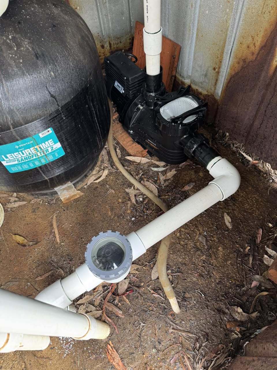 A black pool pump and sand filter with white pipework and a strainer, installed in a tight corner against corrugated iron.
