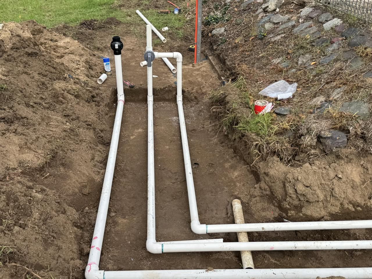 White PVC pipework with valves set out in a wide trench between a lawn edge and a rock retaining bank.
